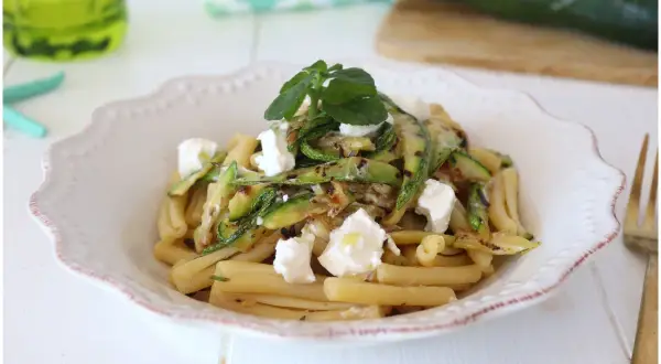 Pasta fredda con zucchine grigliate e cubetti di formaggio fresco, guarnita con erbe aromatiche. Un piatto estivo leggero e gustoso.