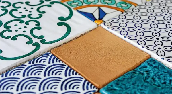 Composizione di mattonelle fai da te con decori geometrici e floreali in blu, verde e giallo, alternate a tasselli color sabbia e turchese.