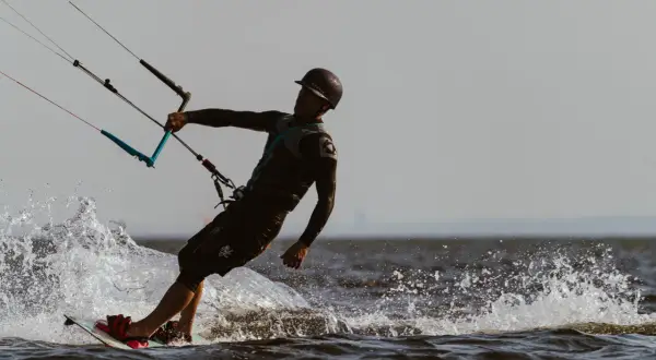 Kitesurfer w kasku i piance, z latawcem i deską, tworzy rozpryski wody na jeziorze.