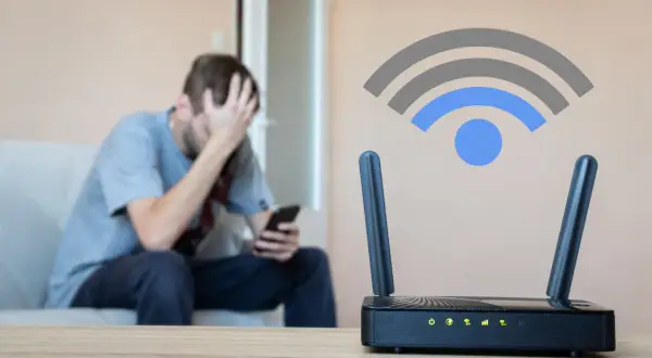 Zmartwiony mężczyzna siedzi na kanapie, trzymając telefon. Obok router Wi-Fi z ikoną sygnału, sugerującą słabą siłę sygnału wifi.