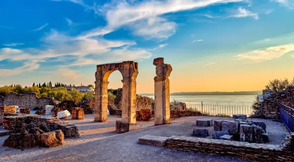 Resti archeologici delle Grotte di Catullo con un arco in pietra e colonne, sullo sfondo il lago e un cielo azzurro al tramonto.