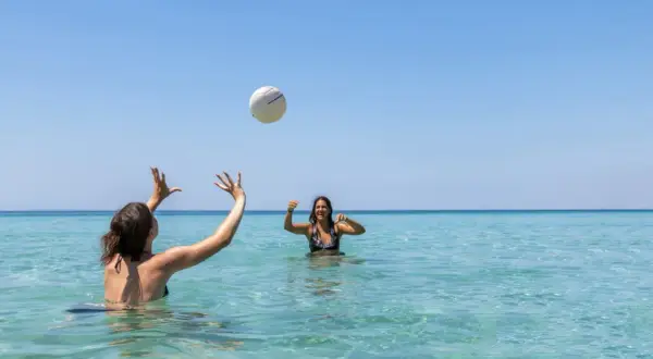 Dos amigas juegan voleibol en el mar turquesa de la playa América, bajo un cielo azul despejado.