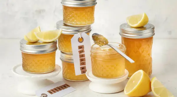 Gläser mit leuchtend gelber Zitronenmarmelade, garniert mit frischen Zitronenspalten. Ein Löffel mit Marmelade zeigt die köstliche Konsistenz, perfekt für ein Zitronenmarmelade Rezept.