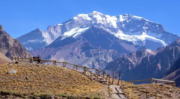Majestatyczny Aconcagua szczyt w Andach, pokryty śniegiem, góruje nad suchym, skalistym krajobrazem. Ścieżka z drewnianą balustradą prowadzi do punktu widokowego.