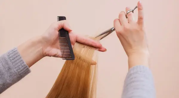 Hände mit Kamm und Schere schneiden blonde Haare. So kannst du kaputte Spitzen selber schneiden und deinem Haar neuen Glanz verleihen.