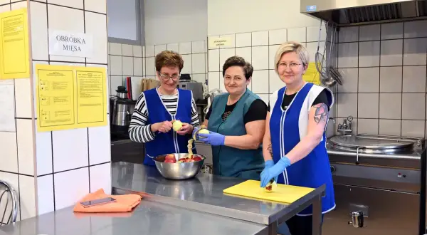 Trzy kobiety w fartuchach obierają jabłka w kuchni. Jedna z nich, intendentka, przygotowuje owoce do dalszej obróbki.
