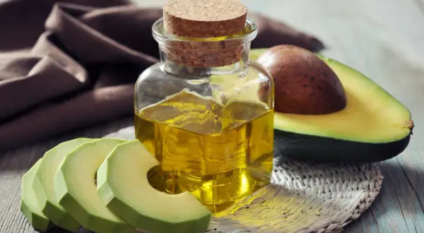 Flasche mit goldenem Persea gratissima oil neben frischen Avocadoscheiben und einer halben Avocado.