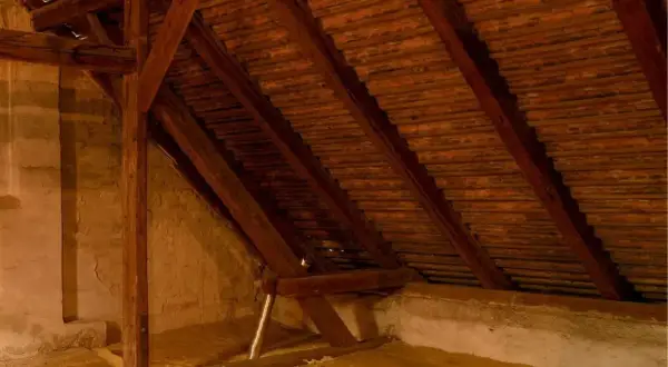 Drewniana konstrukcja poddasza, widoczne belki i dachówki. Na podłodze materiał do ocieplenie poddasza.