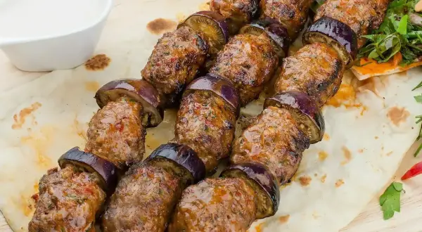 Kofta kebab z bakłażanem na patyku, podany na chlebku z sosem.