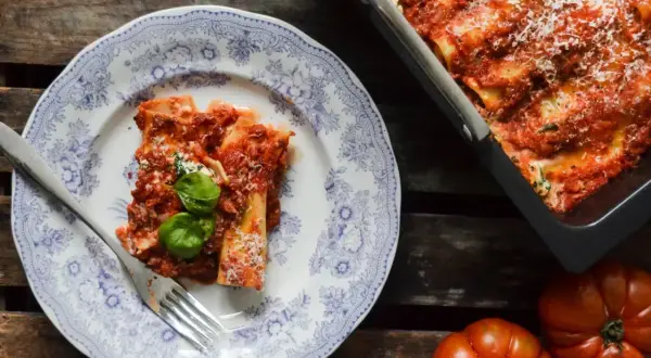 En portion läcker cannelloni med tomatsås och basilika på en vacker tallrik. Bredvid syns en ugnsform med mer cannelloni och färska tomater. Ett perfekt cannelloni recept!