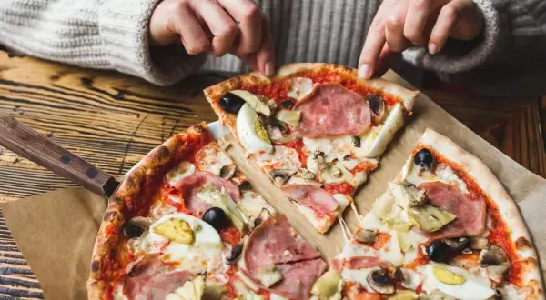 Une personne choisit une part de pizza avec une riche pizza garniture : jambon, œufs, olives, artichauts et champignons.