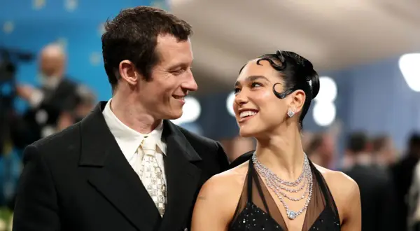 Dua Lipa y su novio, Romain Gavras, sonríen mirándose. Ella luce un vestido negro y joyas deslumbrantes.