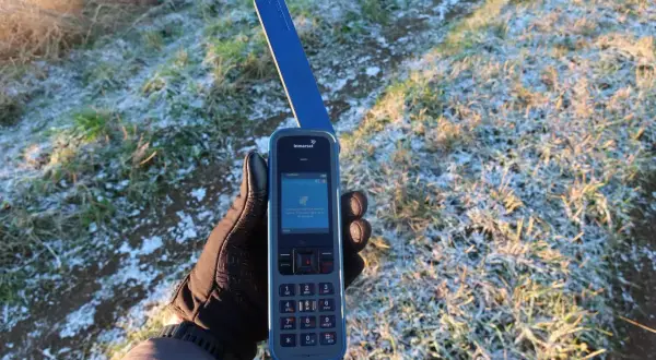 Ręka w rękawiczce trzyma telefon satelitarny Inmarsat IsatPhone, pokazując ekran z informacją o wyszukiwaniu sygnału. Idealne narzędzie, jak sprawdzić prognozę pogody przez telefon satelitarny na wyprawie.