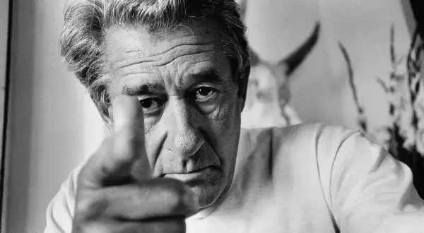 Helmut Newton, z palcem wskazującym w obiektyw, patrzy intensywnie. W tle widać fragmenty jego charakterystycznych prac.