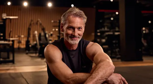 Ein lächelnder Mann mit grauem Haar und Bart sitzt im Fitnessstudio. Er zeigt, dass Muskelaufbau ab 50 möglich ist.