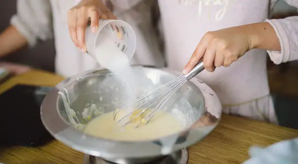 Préparation de la pâte à crêpes : du sucre est versé dans un saladier où une main fouette la préparation.
