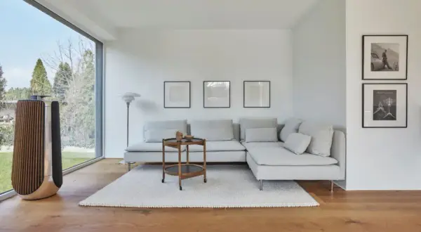 Großes Fenster mit Blick ins Grüne, ein bequemes Ecksofa, ein Beistelltisch und ein Lautsprecher prägen diese moderne Einrichtung.