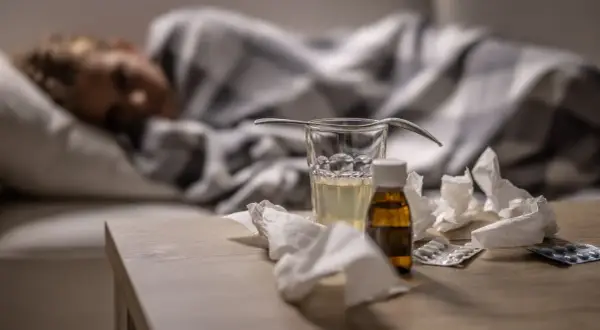 Sjuk person i sängen med medicin, glas vatten och näsdukar på bordet. Viktigt för att minska sjukfrånvaro.