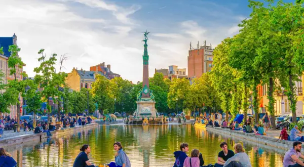 Des gens profitent d'une journée ensoleillée au bord d'un étang, un monument imposant se dresse en arrière-plan. Une belle scène pour visiter Bruxelles.