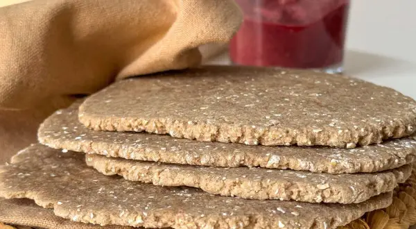 Una pila de pan sin levadura, rústico y con trozos de avena, junto a un vaso de mermelada de frutos rojos y una tela marrón.