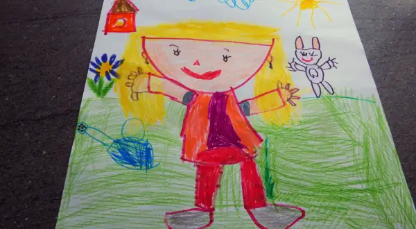 Rysunek dziecka 5 letniego przedstawia uśmiechniętą dziewczynkę w kolorowym ubraniu, domek, słońce, chmurki i królika.