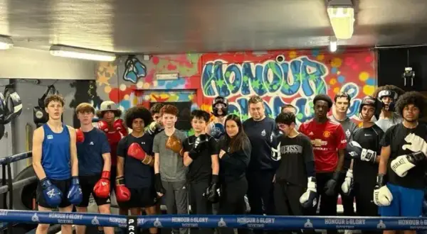 A group of young boxers, some wearing gloves, stand in a boxing ring. One boxer is in the foreground, ready to run.