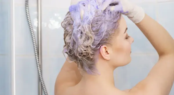 Kobieta w rękawiczkach nakłada fioletowy szampon na blond włosy. Czy szampon koloryzujący niszczy włosy?