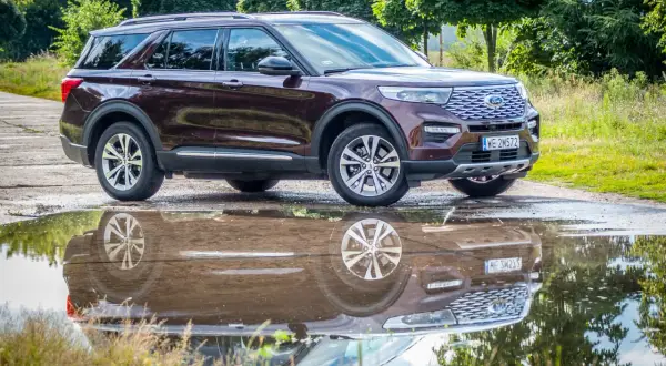 Ciemnobordowy Ford Explorer, jeden z popularnych ford suv modele, odbija się w kałuży po deszczu, otoczony zielenią.
