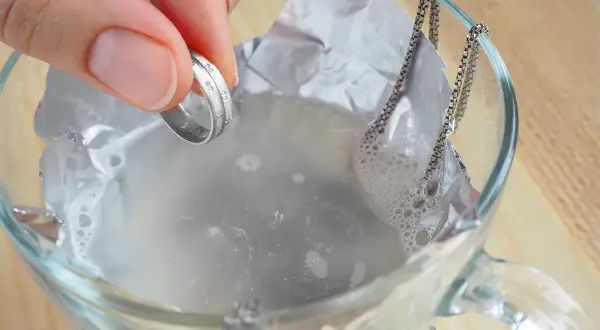 Dłoń trzyma srebrny pierścionek nad szklaną miską z wodą i folią aluminiową, obok wisi srebrny łańcuszek.