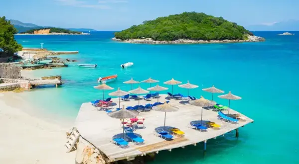 Słoneczny dzień na Ksamil Beach. Drewniany taras z leżakami i parasolami, turkusowa woda i zielona wyspa w tle.