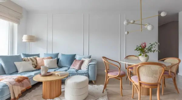 Miękka, błękitna sofa i pudroworóżowe akcenty tworzą przytulną atmosferę. Ciepłe kolory do salonu podkreślają drewniane meble i jasny dywan.