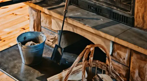 Metalowy kubeł z popiołem i kosz na drewno przy kamiennym kominku. Latem śmierdzi z kominka jak się nie pali, ale przygotowania do jesieni trwają.