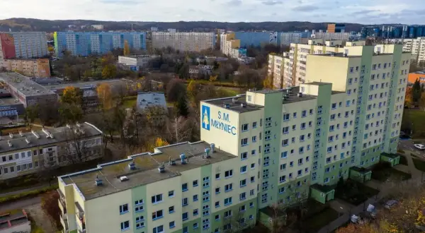 Osiedle z wielkiej płyty S.M. Młyniec, z charakterystycznymi blokami w kolorach zieleni i beżu, otoczone drzewami w jesiennej szacie.