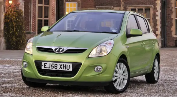 Zielony Hyundai i20 z włączonymi światłami przeciwmgielnymi przednimi, zaparkowany przed ceglanym budynkiem.
