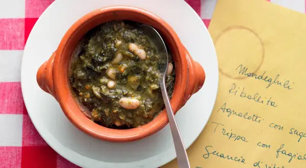 Zuppa di cavolo nero e fagioli in ciotola di terracotta, servita su un piatto bianco con tovaglia a quadri rossi. Accanto, un foglio giallo con un elenco di piatti.