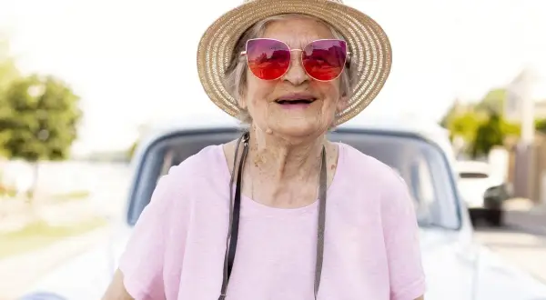Une femme souriante, chapeau de paille et lunettes roses, prête pour partir seule en vacances à 60 ans. Une voiture ancienne en arrière-plan.