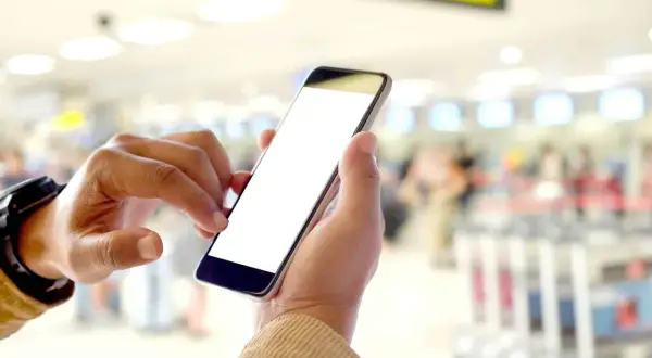 Ręce trzymają smartfon z pustym ekranem. W tle widać zamazany terminal lotniczy, sugerujący, że to idealny moment na **lot odprawa online kiedy** tylko chcesz.