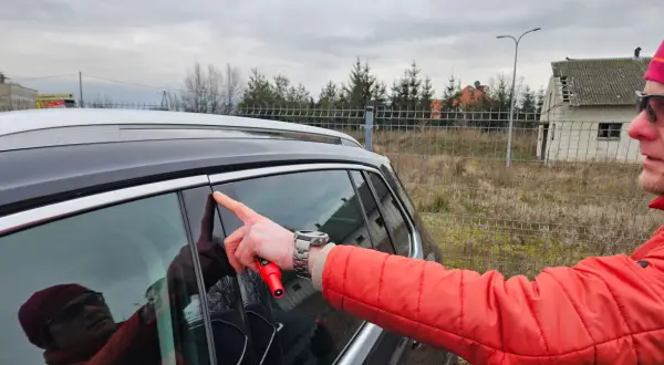 Mężczyzna w czerwonej kurtce wskazuje palcem na drzwi swojego pierwszego samochodu, trzymając w dłoni czerwony przyrząd.