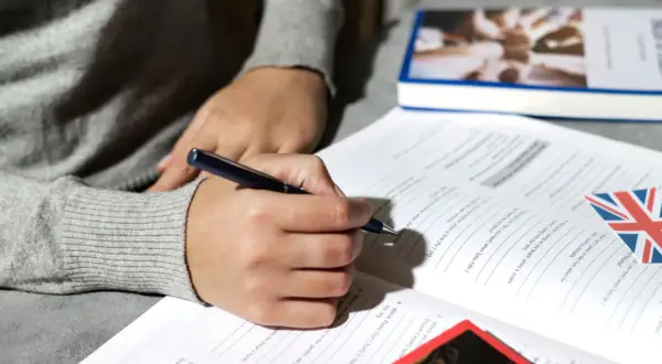 Persona escribiendo en un libro de ejercicios de inglés, con la bandera británica. Preparación para la habilitación lingüística Madrid.