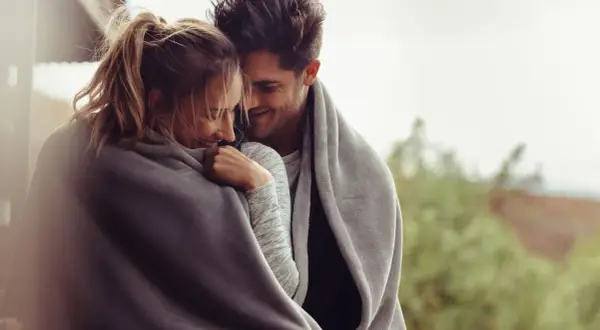 Coppia abbracciata sotto una coperta, sorrisi complici e sguardi teneri, un dolce inizio di una relazione.
