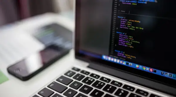 Laptop z kodem na ekranie, obok smartfon. Jak przyspieszyć laptopa? Optymalizacja kodu to klucz.