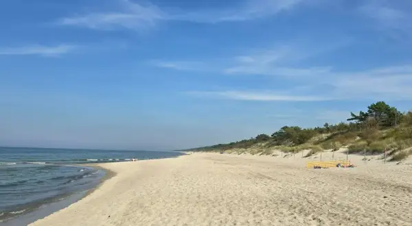Słoneczny dzień nad zalewem kamieńskim. Szeroka, piaszczysta plaża z łagodnymi falami i zielonymi wydmami w tle. Kilka osób odpoczywa na piasku.