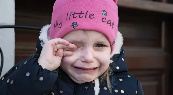 Mała dziewczynka w różowej czapce z napisem "my little cat" płacze, pocierając oko. Wygląda, jakby coś jej wpadło, może opiłek w oku.