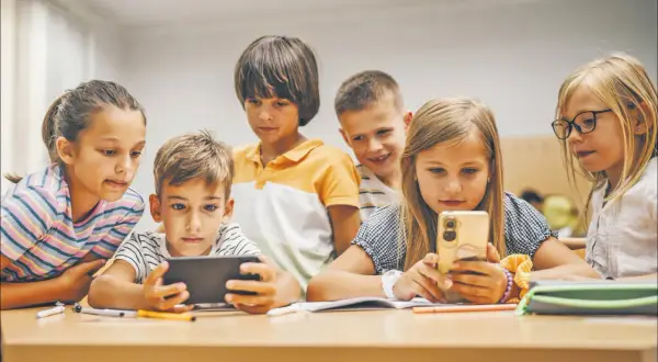 Dzieci skupione na ekranach telefonów i tabletu, uczące się i bawiące razem. Wspólna **socjalizacja** w cyfrowym świecie.