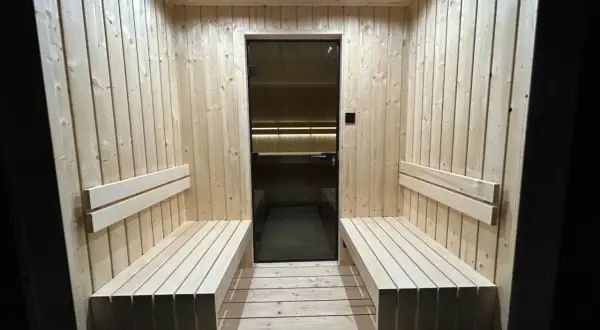 Drewniana sauna z ławami i szklanymi drzwiami. Dyskretna wentylacja w saunie zapewnia komfort.