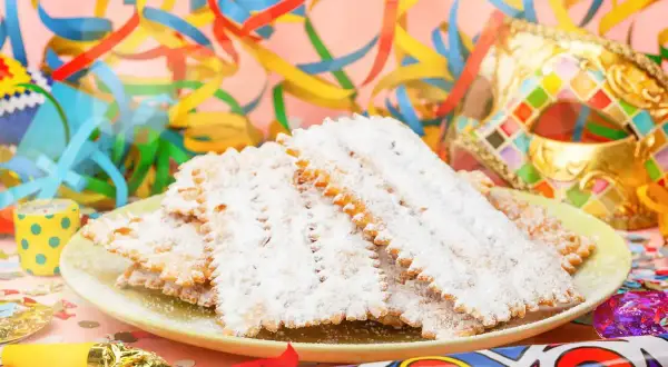 Bugie ricetta originale, dolci fritti cosparsi di zucchero a velo, su un piatto con coriandoli e maschere di Carnevale.
