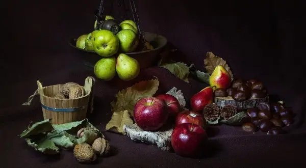 Scatti still life: mele rosse, pere verdi e rosse, noci in un cesto di legno e castagne su un tronco.