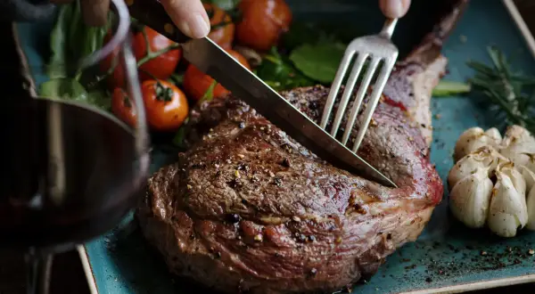 Ein saftiges Lamm-Steak wird mit Messer und Gabel angeschnitten. Dazu passen gerösteter Knoblauch, Kirschtomaten und ein Glas Rotwein. Welcher Wein zu Lamm? Ein kräftiger Rotwein!