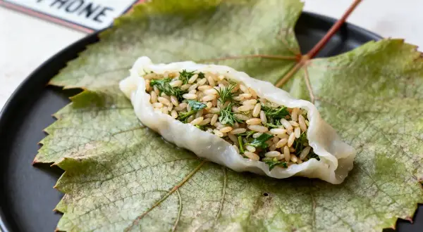 Une feuille de vigne farcie, garnie de riz et d'herbes fraîches, repose sur une grande feuille de vigne verte sur une assiette noire.