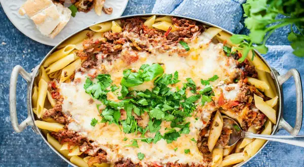 En stor ugnsform med gyllene penne, täckt med köttfärssås och smält ost. En portion pasta al forno är upplagd på en tallrik bredvid.